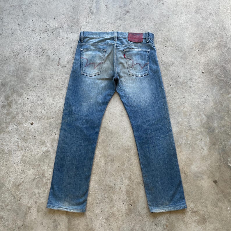 Puede incluir: Vaqueros de mezclilla azul descolorido con un dise&ntilde;o cl&aacute;sico de cinco bolsillos. Los vaqueros presentan un parche de cuero rojo en la parte trasera y costuras distintivas en los bolsillos traseros. Los vaqueros est&aacute;n colocados sobre una superficie de hormig&oacute;n.