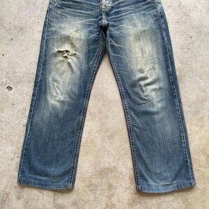 May include: Faded blue denim jeans with a distressed look. The jeans have a relaxed fit, a button and zipper closure, and a worn patch near the left knee. The denim has a vintage wash with visible wear and tear.