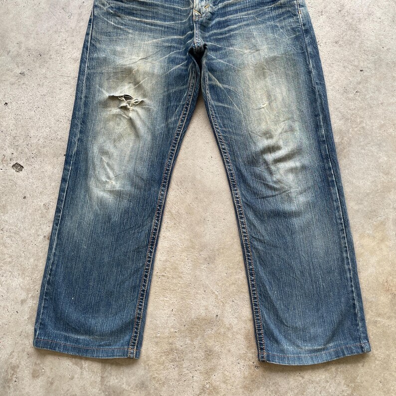 May include: Faded blue denim jeans with a distressed look. The jeans have a relaxed fit, a button and zipper closure, and a worn patch near the left knee. The denim has a vintage wash with visible wear and tear.