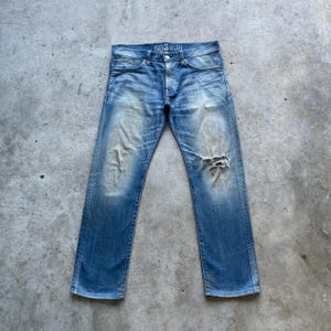 Puede incluir: Vaqueros de mezclilla azul claro con un aspecto desgastado. Los vaqueros tienen un lavado descolorido, una rodilla rasgada y el texto "503 SLIM" en el bolsillo trasero. Los vaqueros est&aacute;n colocados planos sobre una superficie de hormig&oacute;n.