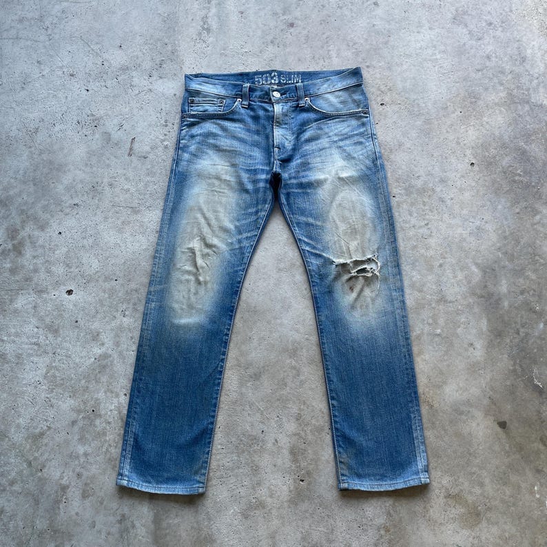 Puede incluir: Vaqueros de mezclilla azul claro con un aspecto desgastado. Los vaqueros tienen un lavado descolorido, una rodilla rasgada y el texto "503 SLIM" en el bolsillo trasero. Los vaqueros est&aacute;n colocados planos sobre una superficie de hormig&oacute;n.