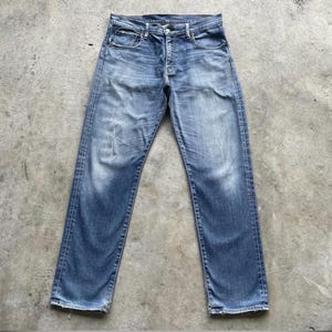 May include: Faded blue denim jeans with a classic five-pocket design. The jeans have a button closure and a straight leg fit. The denim shows signs of wear and fading, giving them a vintage look. The jeans are laid flat on a concrete surface.