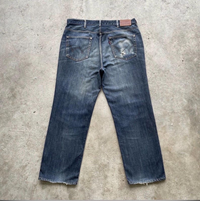 K&ouml;nnte beinhalten: Verblasste blaue Jeans mit klassischem F&uuml;nf-Taschen-Design. Die Jeans haben ein abgenutztes Aussehen mit abgenutzten Details, einschlie&szlig;lich eines kleinen Risses an der Ges&auml;&szlig;tasche. Ein brauner Lederaufn&auml;her ist am Bund sichtbar.