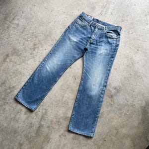 May include: Light blue denim jeans with a classic five-pocket design. The jeans have a straight leg cut and show signs of wear, with faded areas and subtle distressing. The jeans are laid flat on a concrete surface.