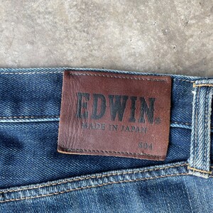 Puede incluir: Primer plano de vaqueros de mezclilla azul con un parche de cuero marr&oacute;n. El parche presenta el nombre de la marca "EDWIN" en relieve, junto con "MADE IN JAPAN" y el n&uacute;mero "304". Los vaqueros tienen costuras visibles.