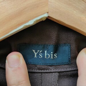 May include: Close-up of a brown fabric with a dark blue label. The label reads "Y's bis" in a silver font. The fabric is held by a wooden hanger.