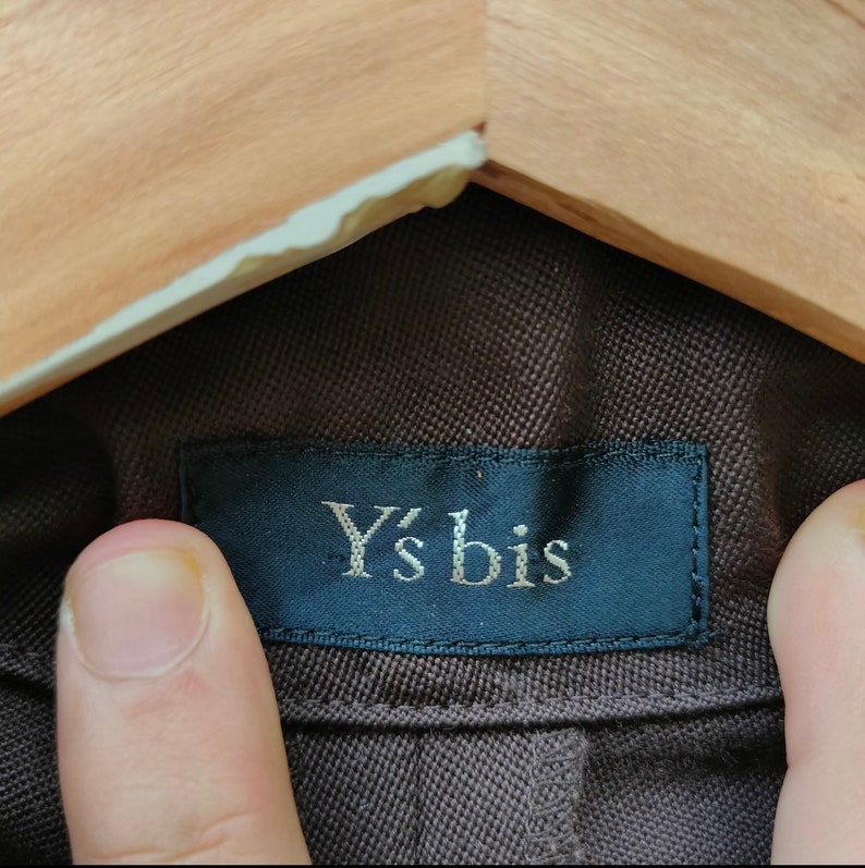 May include: Close-up of a brown fabric with a dark blue label. The label reads "Y's bis" in a silver font. The fabric is held by a wooden hanger.