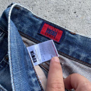 Pu&ograve; includere: Primo piano di jeans blu con una toppa rettangolare rossa che recita "JEANS 503 EDWIN". Un'etichetta bianca con testo nero &egrave; visibile, compreso il marchio "EDWIN" e informazioni sulla taglia.