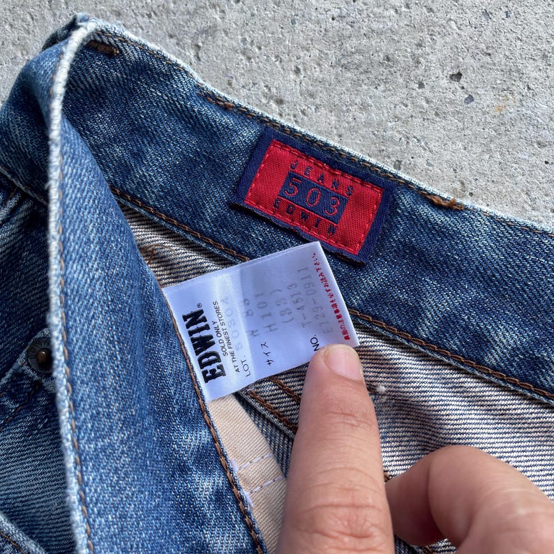 Pu&ograve; includere: Primo piano di jeans blu con una toppa rettangolare rossa che recita "JEANS 503 EDWIN". Un'etichetta bianca con testo nero &egrave; visibile, compreso il marchio "EDWIN" e informazioni sulla taglia.