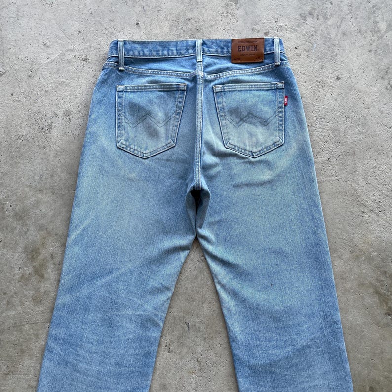 May include: Light blue denim jeans with a classic five-pocket design. The jeans feature a faded wash and a straight leg cut. The back pockets have a unique stitching pattern. A brown leather patch with the brand name "EDWIN" is visible.
