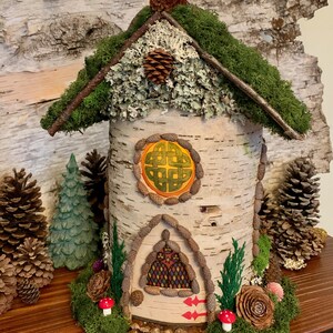 Könnte beinhalten: Ein Miniatur-Feenhaus aus Birkenrinde mit einem Moosdach und einem keltischen Knotendesign am Fenster. Das Haus ist umgeben von Tannenzapfen, Moos und anderen natürlichen Elementen.