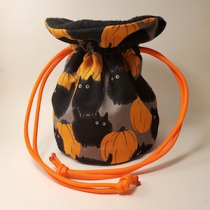 May include: A drawstring pouch featuring a Halloween-themed pattern. The fabric displays black cats and orange pumpkins on a gray background. The pouch has an orange drawstring closure and a black rim.