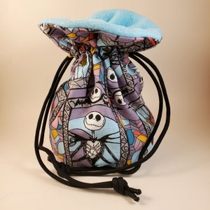 Puede incluir: Una bolsa de dados de tela azul y morada con un patrón de vitral que presenta a Jack Skellington de Pesadilla antes de Navidad. La bolsa tiene un cordón de ajuste negro y un forro de vellón azul.