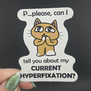 May include: A die-cut sticker featuring a tan cartoon cat with large, sparkling eyes and pink cheeks, holding its paws together. The text above reads P...please, can I and the text below reads tell you about my CURRENT HYPERFIXATION?