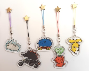 May include: Six colorful phone charms with different cartoon characters. The charms are attached to gold star-shaped charms with strings in various colors. The characters include a white hedgehog, a blue hedgehog on a skateboard, a black hedgehog on a motorcycle, a red hedgehog holding a green diamond, a yellow fox with a propeller, and a white fox with a propeller.