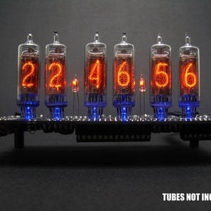 Pode incluir: Um suporte de metal preto com seis tubos Nixie de vidro transparente que exibem a hora 22:46:56. Os tubos são iluminados com números laranja e um brilho azul ao redor da base. "TUBES NOT INCLUDED" está impresso abaixo do suporte.