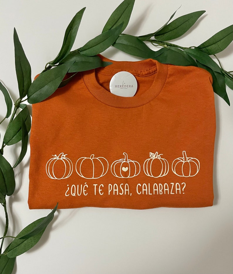Puede incluir: Una camiseta naranja con un gr&aacute;fico blanco de cinco calabazas y el texto "&iquest;Qu&eacute; te pasa, calabaza?"