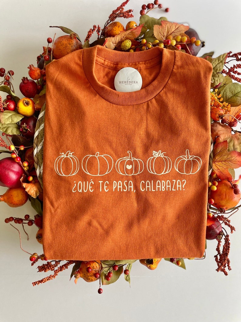 May include: An orange t-shirt with a white graphic of five pumpkins and the text '¿Qué te pasa, calabaza?' The t-shirt has a white circle label with the text 'HEREDERA' on it.