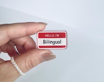 Puede incluir: Una etiqueta rectangular con borde rojo e interior blanco. El texto "HELLO, I'M" está en la parte superior, y "Bilingual" está escrito en negrita, sin serifa, debajo. La etiqueta se sostiene entre los dedos.
