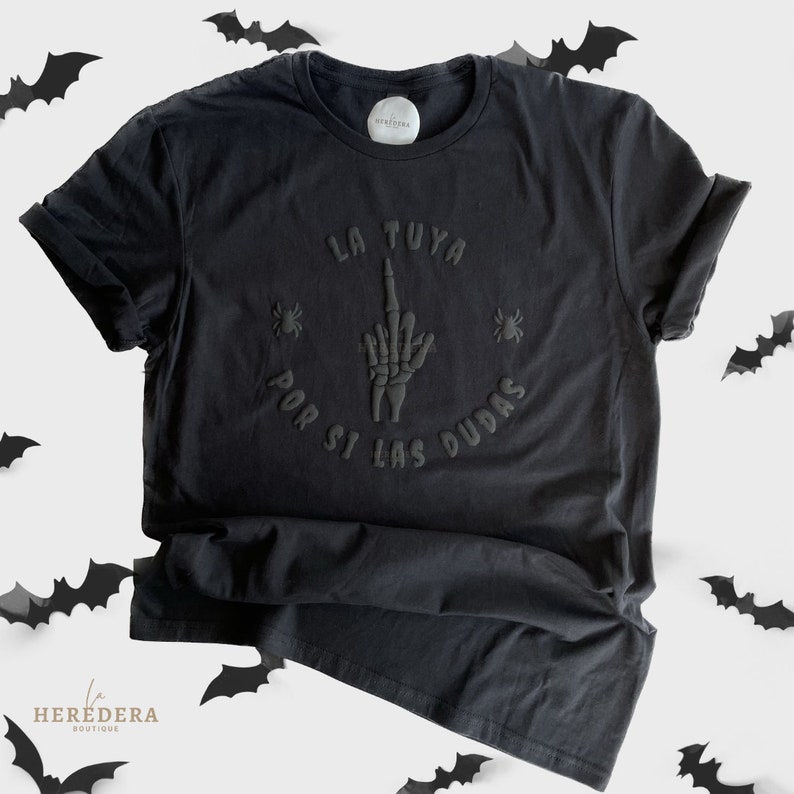 Puede incluir: Camiseta negra con una mano de esqueleto negra haciendo el dedo medio y el texto "La Tuya Por Si Las Dudas" en espa&ntilde;ol.
