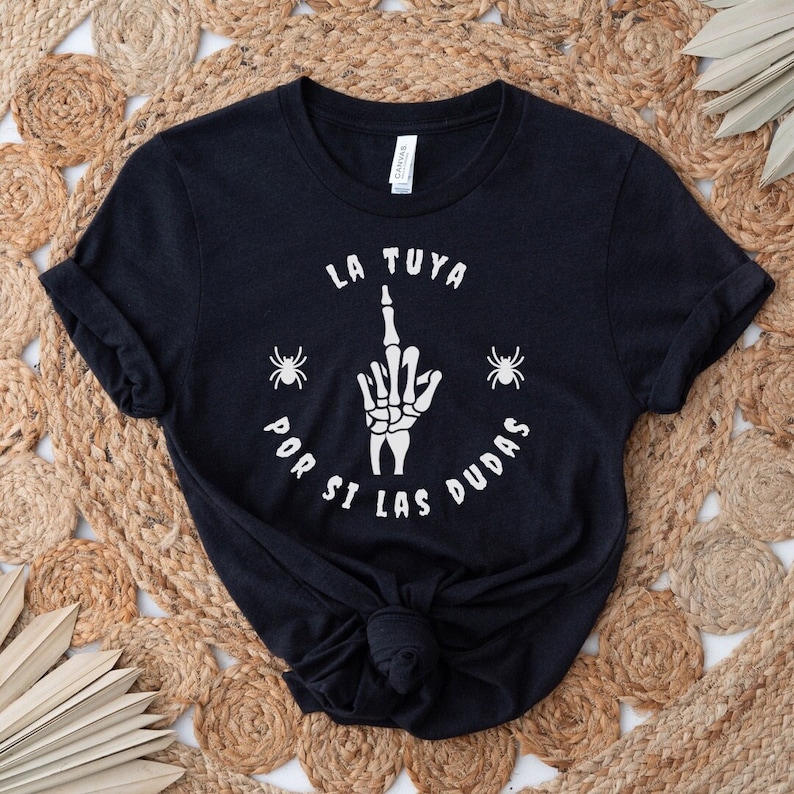 Puede incluir: Camiseta negra con una mano de esqueleto blanca haciendo el dedo medio, rodeada del texto "LA TUYA POR SI LAS DUDAS" y dos siluetas de ara&ntilde;a blancas.