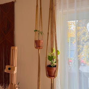 Op de afbeelding: Twee beige macramé plantenhangers gemaakt van geknoopt koord, elk met een terracotta pot met een kleine groene plant. De hangers zijn voorzien van verticale koorden en decoratieve knopen en hangen op verschillende hoogtes aan het plafond.