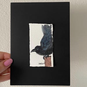 May include: A watercolor painting of a black crow perched on a brown post, set against a white background. The artwork is mounted on a black rectangular card. The artist's signature is visible on the artwork.