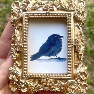 May include: A small, ornate gold frame holds a watercolor painting of a dark blue bird with its beak open. The frame has intricate scrollwork and a glossy finish, creating a vintage aesthetic. The bird is painted on a white background.