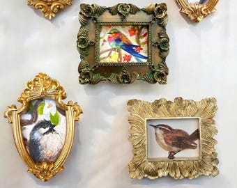 May include: Six ornate gold frames with various bird paintings inside. The frames are all different shapes and sizes, and the birds are depicted in a variety of poses. The frames are arranged in a row on a white wall.