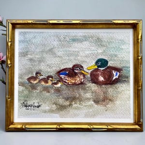 May include: Watercolor painting of a family of ducks in a gold bamboo-style frame. The painting depicts a male duck with a green head and yellow beak, two female ducks, and three ducklings. The ducks are brown and white, swimming in a body of water.