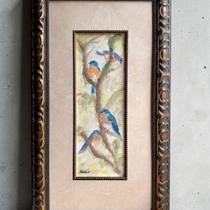 Può includere: Un dipinto verticale ad acquerello raffigurante tre uccelli azzurri appollaiati su rami d'albero, inserito in un passe-partout rettangolare beige e una cornice decorata marrone scuro con riflessi dorati. Firma presente nell'angolo in basso a sinistra.