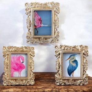 Op de afbeelding: Drie ingelijste vogelkunstprints. De lijsten zijn goudkleurig met gedetailleerde gravures. De prints tonen een roze flamingo, een blauwe reiger en een roze lepelaar in vlucht, tegen een blauwe achtergrond. De lijsten worden op een houten oppervlak weergegeven.
