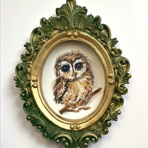 May include: An oval-shaped framed artwork featuring a watercolor painting of a brown owl perched on a branch. The frame has a gold inner border and a green, ornate outer frame. The artwork is set against a white background.