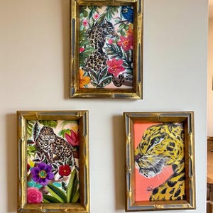 May include: Three framed paintings with gold bamboo-style frames. The paintings depict leopards in lush, colorful floral settings. One painting features a leopard with a blue bird. The other two paintings show leopard faces.