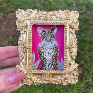 May include: A small, ornate gold frame surrounds a painting of a leopard against a bright pink background. The leopard has blue eyes and a spotted coat. Green and blue leaves are at the bottom of the painting.