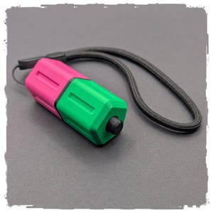 Könnte beinhalten: Kleines, farbenfrohes Gerät mit schwarzem Knopf und schwarzer Handschlaufe. Das Gerät ist in zwei Abschnitte unterteilt: einen rosa Abschnitt und einen grünen Abschnitt. Die Handschlaufe besteht aus einem schwarzen, gewebten Material.