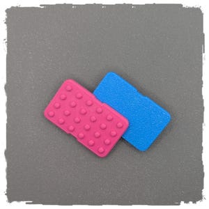 Könnte beinhalten: Zwei kleine, rechteckige Kunststoffartikel. Einer ist leuchtend rosa mit gleichmäßig verteilten, erhabenen kreisförmigen Erhebungen. Der andere ist blau mit einer strukturierten Oberfläche. Beide liegen auf einer grauen Oberfläche.