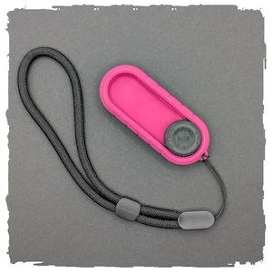 Könnte beinhalten: Ein leuchtend pinkfarbenes, oval geformtes Gerät mit strukturiertem Rand und einer dunkelgrauen kreisförmigen Komponente. Ein schwarzes gewebtes Handgelenkband ist befestigt. Das Gerät befindet sich vor einem grauen Hintergrund.