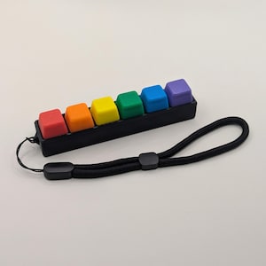 Könnte beinhalten: Ein schwarzes, rechteckiges Makro-Tastenfeld mit sechs quadratischen Tasten in einer Regenbogenabfolge aus Rot, Orange, Gelb, Grün, Blau und Violett. Das Gerät verfügt über eine schwarze Nylon-Handschlaufe mit einem verstellbaren Kunststoffschieber.