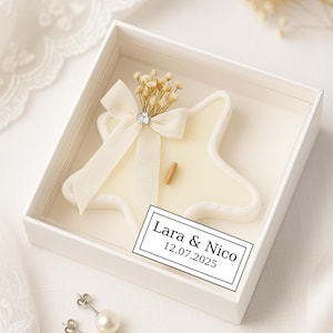 Può includere: Una candela a forma di stella, color avorio, con uno stoppino in legno, presentata in una scatola bianca. La candela è adornata con un fiocco color crema, un piccolo cristallo e fiori secchi. L'etichetta recita "Lara & Nico 12.07.2025".