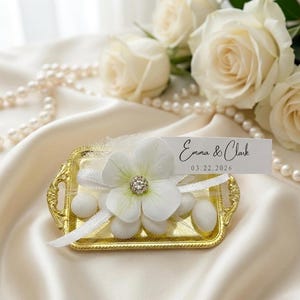 May include: A small rectangular gold-toned metal tray with ornate handles, holding white sugared almonds, a white fabric flower with a central rhinestone, and a white rectangular tag printed with the text Emma & Clark 03.22.2026.