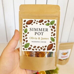 May include: A brown paper bag labeled "Simmer Pot, Olivia & James, Wedding Favor." The label features coffee beans, cocoa beans, and mint leaves. The bag contains dried orange slices and spices. A second bag is visible in the background.