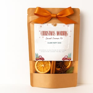 May include: A brown paper pouch with a burnt orange ribbon bow at the top. The label reads CHRISTMAS MORNING Spiced Simmer Kit CLARK PARTY 2025 with illustrations of red plaid trucks. A clear window reveals dried orange slices, cinnamon sticks, and star anise.