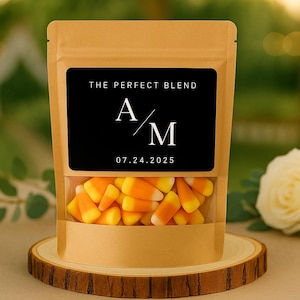 May include: A kraft paper bag with a clear window filled with candy corn. The bag has a black label with the text "THE PERFECT BLEND", initials "A/M", and the date "07.24.2025". The bag sits on a wooden slice.