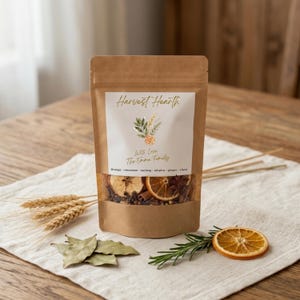 May include: A brown kraft paper pouch containing dried spices and fruit slices. The label reads Harvest Hearth, With Love The Emma Family, and lists orange, cinnamon, nutmeg, allspice, ginger, clove. A clear window reveals the contents inside.
