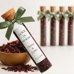 May include: A glass tube filled with dried tea leaves, labelled "Love is Brewing" with names and a date, tied with a green ribbon. A wooden bowl of tea leaves and several similar tubes are in the background.
