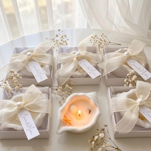 Könnte beinhalten: Weiß geformte Muschelschalen-Kerzen mit kleinen Muscheln und einem Seestern im Inneren, präsentiert in transparenten Boxen. Jede Box ist mit einem cremefarbenen Band und einem Etikett mit der Aufschrift "Emma & Matia" versehen.