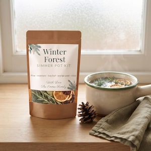 May include: A brown paper pouch labelled Winter Forest Simmer Pot Kit with text listing pine, rosemary, bay leaf, orange peel, and clove. Beside the pouch sits a small pinecone and a ceramic mug filled with steaming water, orange slices, and herbs.