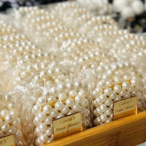 May include: Close-up of several small bags filled with white pearl necklaces. Each bag has a gold label that reads "Laura's Bridal Shower" and the date "10-25". The bags are arranged in a wooden tray.