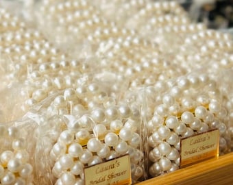 May include: Close-up of several small bags filled with white pearl necklaces. Each bag has a gold label that reads "Laura's Bridal Shower" and the date "10-25". The bags are arranged in a wooden tray.